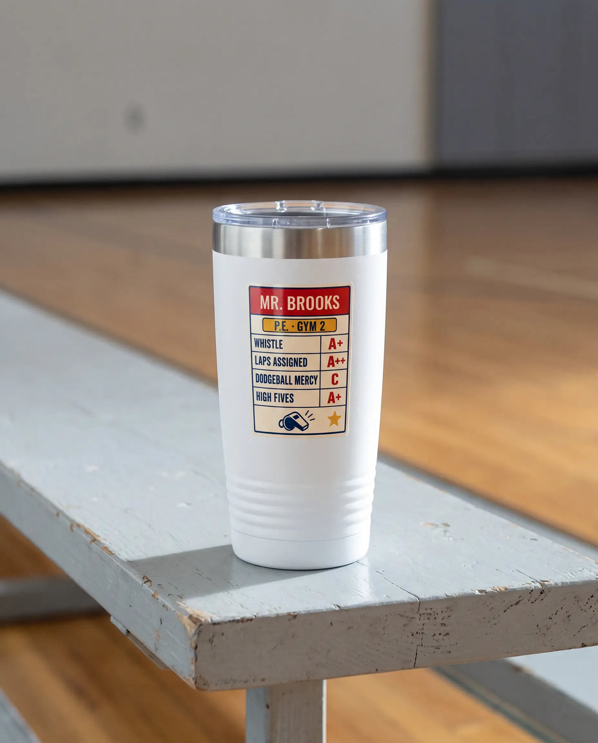 Teacher Report Card, P.E. badge on a white 20oz tumbler personalized MR. BROOKS, P.E. - GYM 2, the pale gray bleacher bench of a school gym