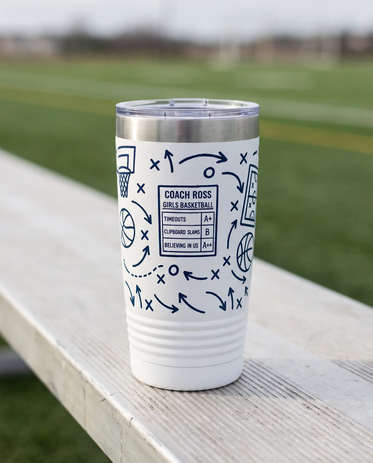 Coach Chalk Talk, Basketball wrap on a white 20oz tumbler personalized COACH ROSS, GIRLS BASKETBALL, three-quarter view showing the ribbed lower wall, a sideline bench beside an empty green sports field