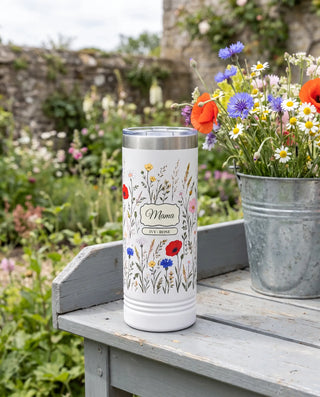 Wildflower Meadow wrap on a white 22oz Skinny tumbler personalized Mama, IVY - ROSE, a gray-painted potting bench in a cottage garden