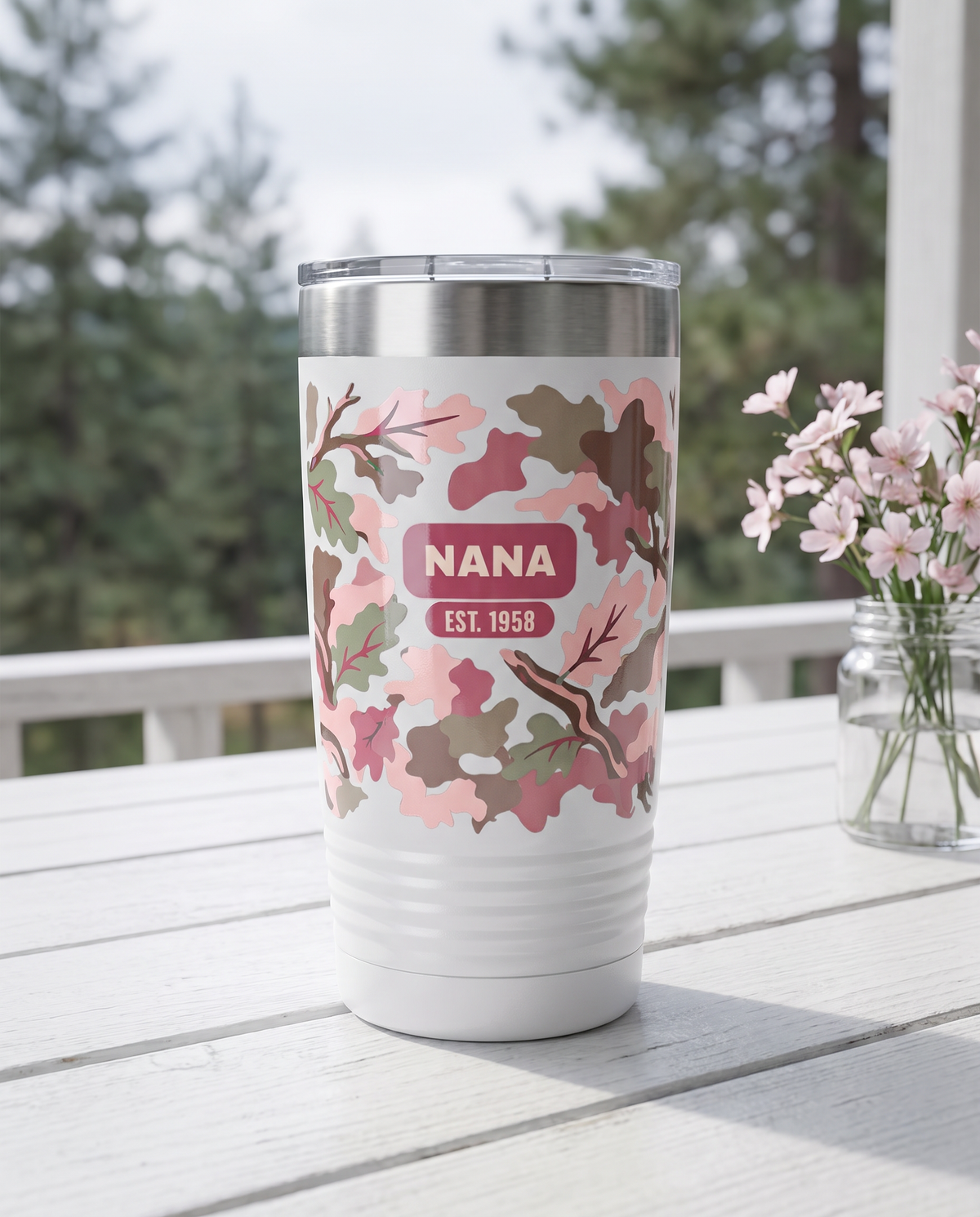 Blush Camo & Bows, Est. Year wrap on a white 20oz tumbler personalized NANA, EST. 1958, a white-painted table on a cabin porch with pale pink wildflowers