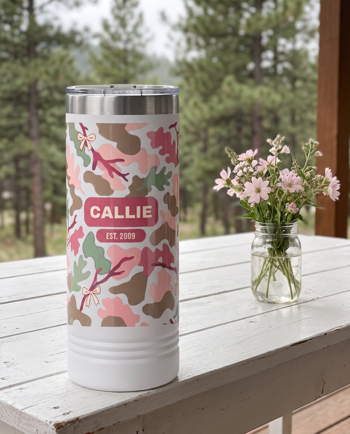 Blush Camo & Bows, Est. Year wrap on a white 22oz Skinny tumbler personalized CALLIE, EST. 2009, a white-painted table on a cabin porch with pale pink wildflowers