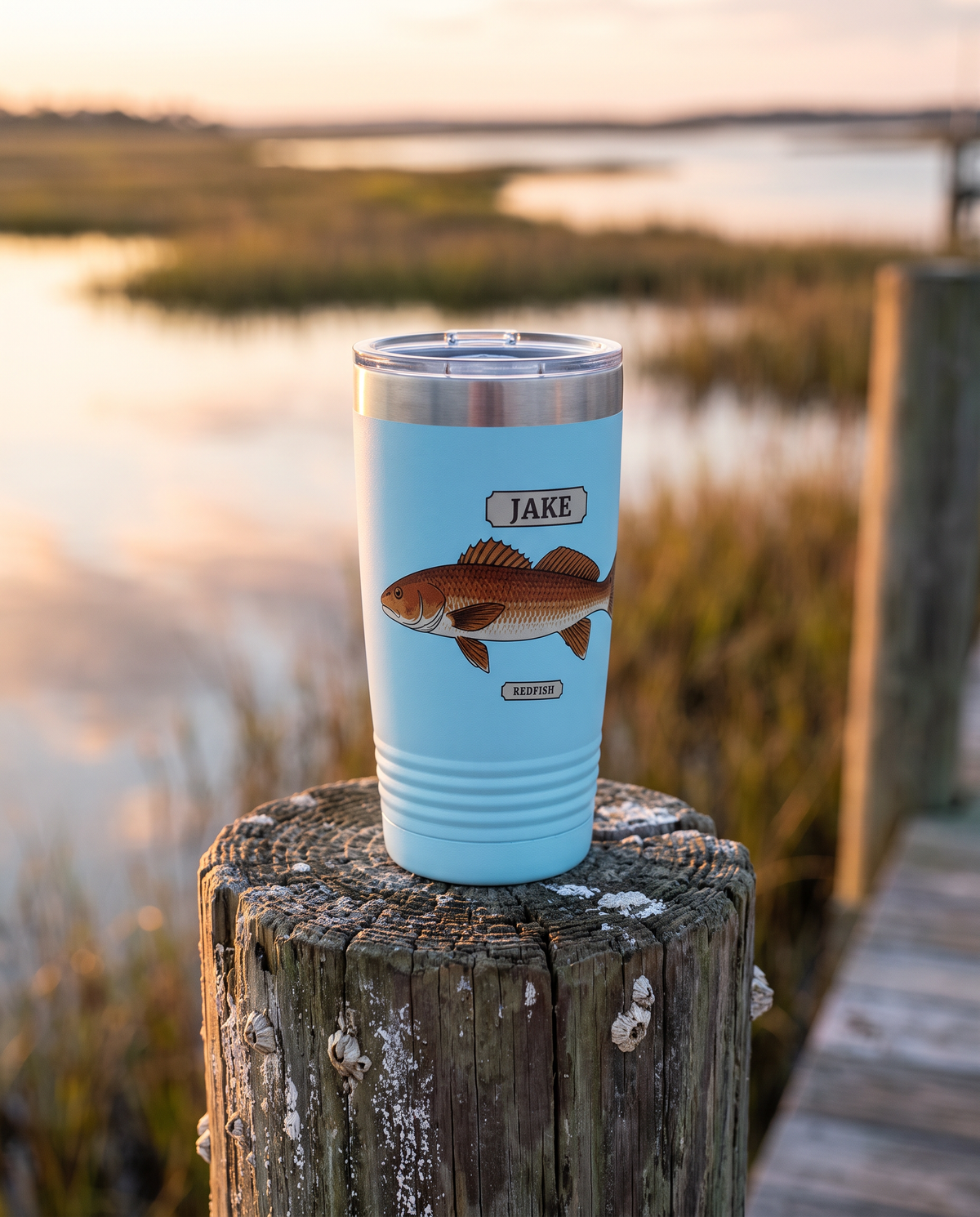 20oz Ringneck tumbler in light blue with the Field Guide Redfish design and a name plate, standing on a dock piling at sunrise