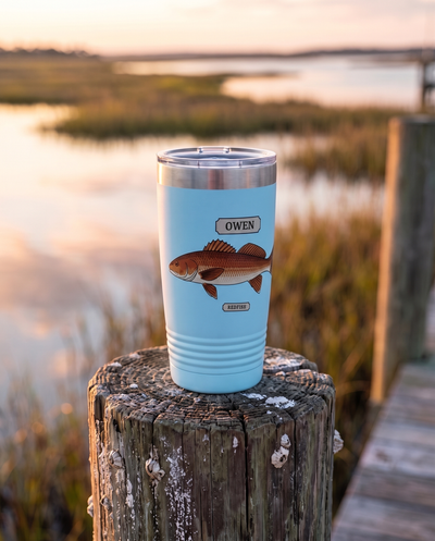 20oz Ringneck tumbler in light blue with the Field Guide Redfish design and a name plate, standing on a dock piling at sunrise, name OWEN