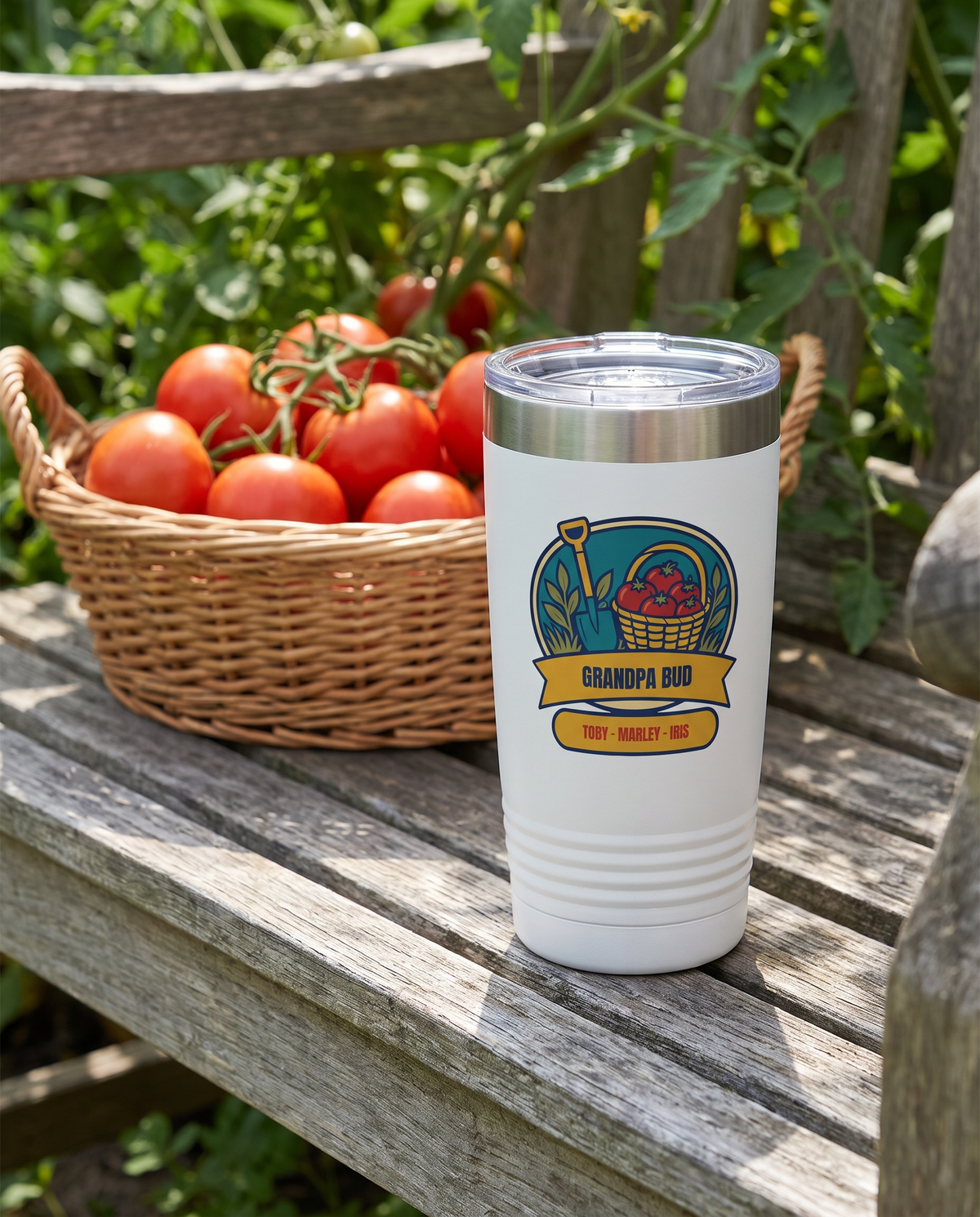 Tomato Harvest Badge, a garden badge in teal, red, gold, cream and olive, UV printed on the front of a 20oz Ringneck tumbler in White, sample name GRANDPA BUD, sits on a garden bench in a vegetable garden, a basket of ripe tomatoes and green vines far behind