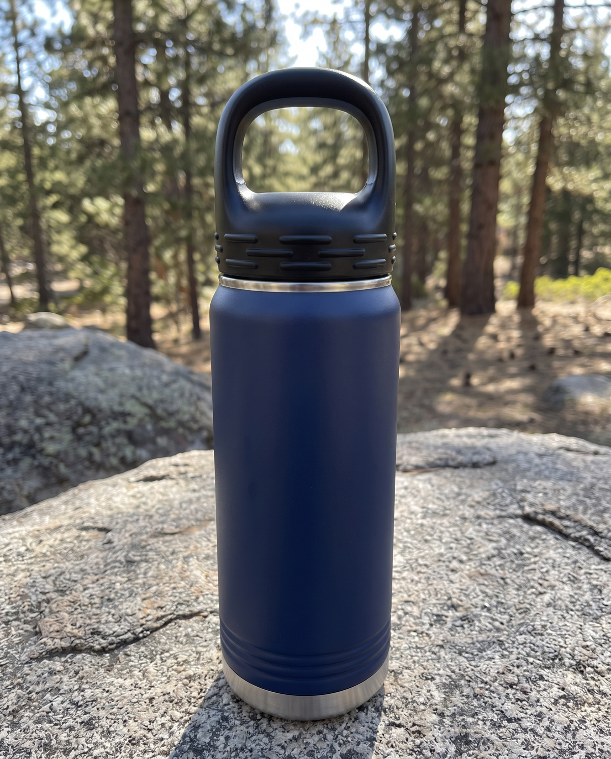 Black lug lid with carry loop on a navy 20oz water bottle, on a granite boulder by a pine trail