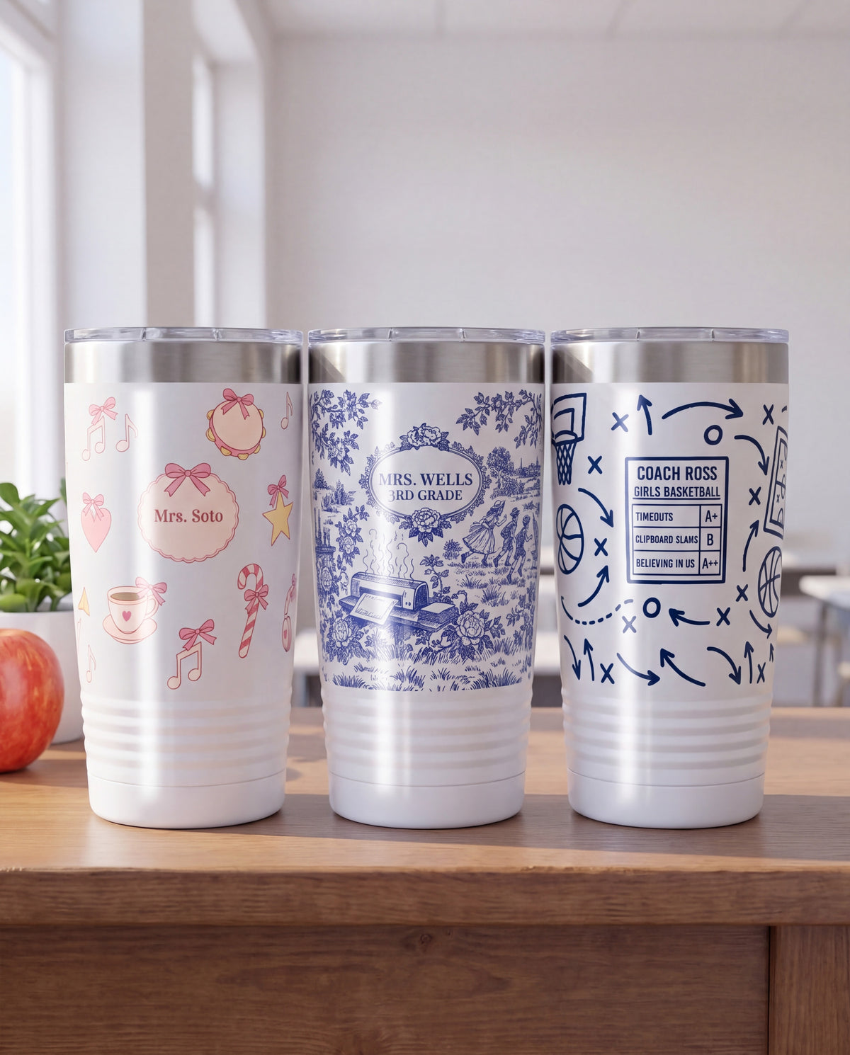 Three personalized white 20oz ringneck tumblers on a wooden teacher's desk in a sunny classroom: a pink bow-tied music room wrap with notes, a tambourine, a teacup and a candy cane reading Mrs. Soto, a blue and white teacher life toile wrap reading MRS. WELLS and 3RD GRADE, and a navy coach chalk talk basketball wrap reading COACH ROSS, GIRLS BASKETBALL, beside a red apple and a potted plant