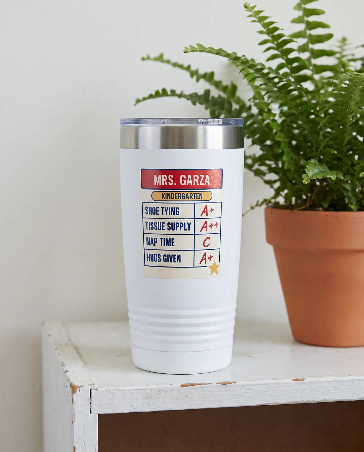 Teacher Report Card, Kindergarten badge on a white 20oz tumbler personalized MRS. GARZA, KINDERGARTEN, a white-painted classroom shelf beside a potted fern