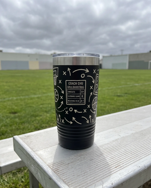 Coach Chalk Talk, Basketball wrap on a black 20oz tumbler personalized COACH CHU, GIRLS BASKETBALL, a white bench outdoors beside an empty green sports field on an overcast day