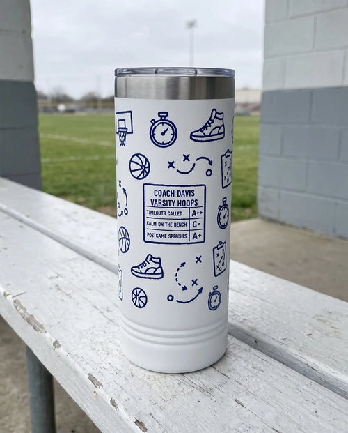 Coach Chalk Talk, Basketball Plays wrap on a white 22oz Skinny tumbler personalized COACH DAVIS, VARSITY HOOPS, a white bench outdoors beside an empty green sports field on an overcast day