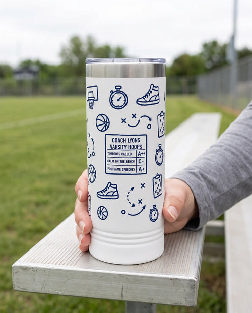 Coach Chalk Talk, Basketball Plays wrap on a white 22oz Skinny tumbler personalized COACH LYONS, VARSITY HOOPS, held in one hand, a sideline bench beside an empty green sports field