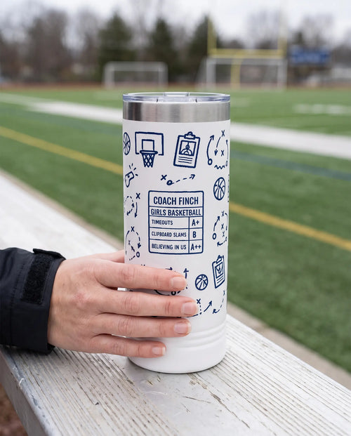 Coach Chalk Talk, Basketball wrap on a white 22oz Skinny tumbler personalized COACH FINCH, GIRLS BASKETBALL, held in one hand, a sideline bench beside an empty green sports field
