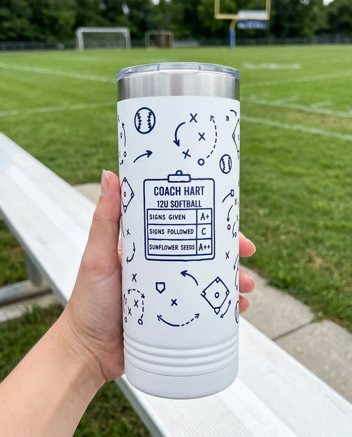 Coach Chalk Talk, Softball wrap on a white 22oz Skinny tumbler personalized COACH HART, 12U SOFTBALL, held in one hand, a sideline bench beside an empty green sports field