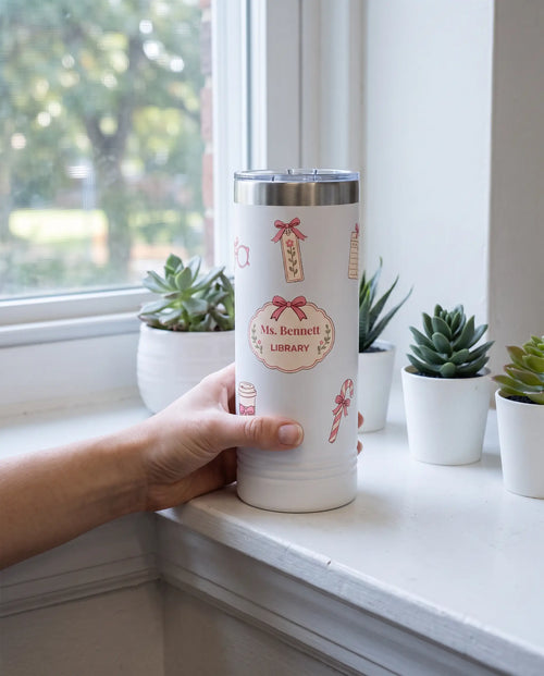 Bow Toss, Librarian wrap on a white 22oz Skinny tumbler personalized Ms. Bennett, LIBRARY, held in one hand, a classroom windowsill in morning light