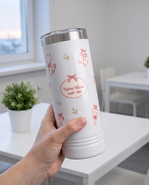 Bow Toss, L&D Nurse wrap on a white 22oz Skinny tumbler personalized Nurse Shaw, L&D - RN, held in one hand, a white hospital break-room table by a window at dawn