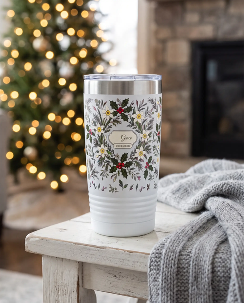 December Birth Flowers wrap on a white 20oz tumbler personalized Grace, three-quarter view showing the ribbed lower wall, a fireside side table with a knit throw