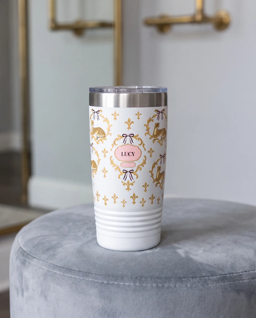 Cheetah Damask wrap on a white 20oz tumbler personalized LUCY, three-quarter view showing the ribbed lower wall, a velvet ottoman in a boutique dressing room