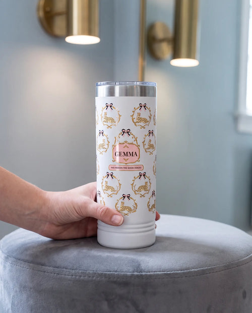 Cheetah Damask, Good Snacks wrap on a white 22oz Skinny tumbler personalized GEMMA, held in one hand, a velvet ottoman in a boutique dressing room