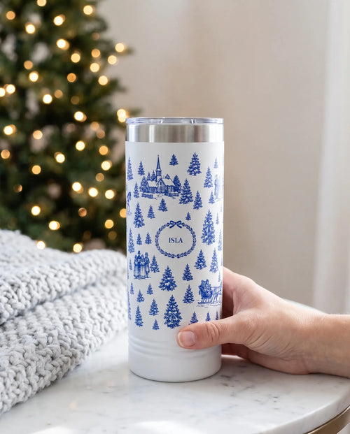 Christmas Toile wrap on a white 22oz Skinny tumbler personalized ISLA, held in one hand, a fireside side table with a knit throw