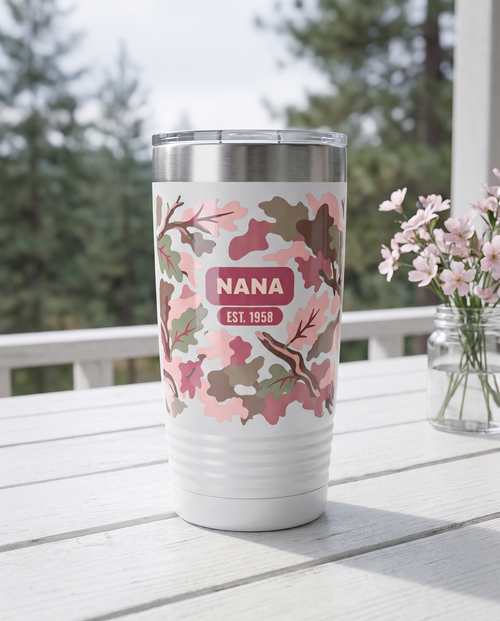 Blush Camo & Bows, Est. Year wrap on a white 20oz tumbler personalized NANA, EST. 1958, a white-painted table on a cabin porch with pale pink wildflowers