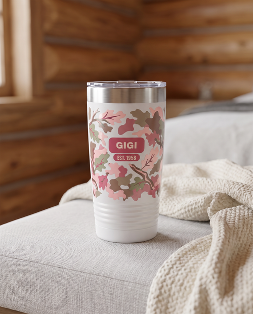 Blush Camo & Bows, Est. Year wrap on a white 20oz tumbler personalized GIGI, EST. 1958, three-quarter view showing the ribbed lower wall, a grey linen bench in a cozy cabin bedroom