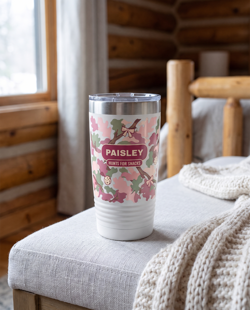 Blush Camo & Bows, Hunts for Snacks wrap on a white 20oz tumbler personalized PAISLEY, HUNTS FOR SNACKS, three-quarter view showing the ribbed lower wall, a grey linen bench in a cozy cabin bedroom