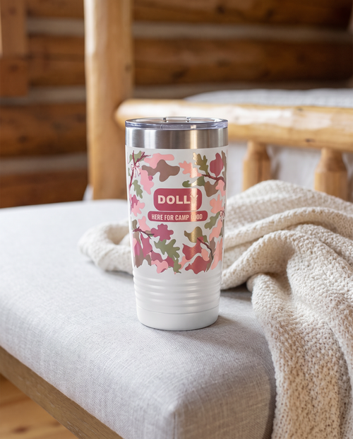 Blush Camo & Bows, Here for Camp Food wrap on a white 20oz tumbler personalized DOLLY, HERE FOR CAMP FOOD, three-quarter view showing the ribbed lower wall, a grey linen bench in a cozy cabin bedroom