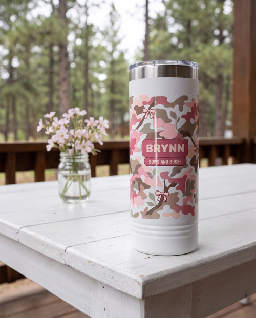 Blush Camo & Bows, Bows and Bucks wrap on a white 22oz Skinny tumbler personalized BRYNN, BOWS AND BUCKS, a white-painted table on a cabin porch with pale pink wildflowers