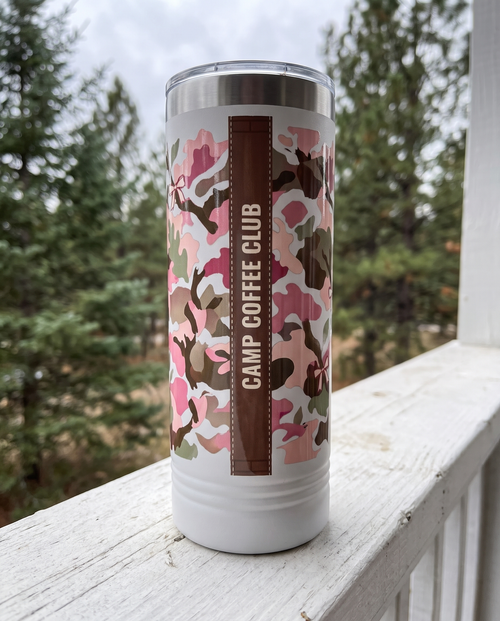 Back of a white 22oz Skinny tumbler with the Blush Camo & Bows, Bows and Bucks wrap, the band reading CAMP COFFEE CLUB, a white-painted cabin porch rail with pines behind