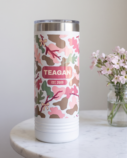 Blush Camo & Bows, Est. Year wrap on a white 22oz Skinny tumbler personalized TEAGAN, EST. 2009, a white marble side table with a jar of pale pink wildflowers
