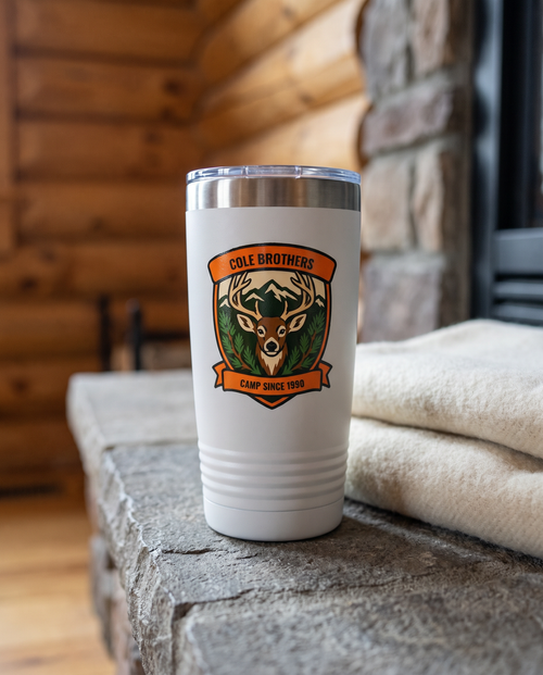 Deer Camp Patch, Buck Shield badge on a white 20oz tumbler personalized COLE BROTHERS, CAMP SINCE 1990, a stone hearth ledge in a log cabin