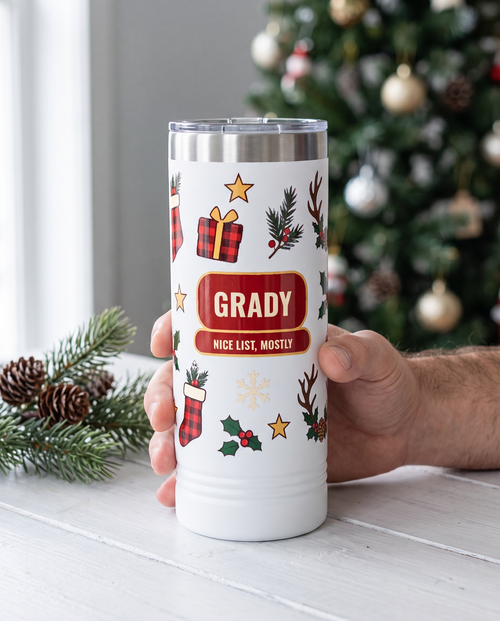 Deer Camp Christmas, Nice List Mostly wrap on a white 22oz Skinny tumbler personalized GRADY, NICE LIST, MOSTLY, held in one hand, a white-painted cabin table at Christmas