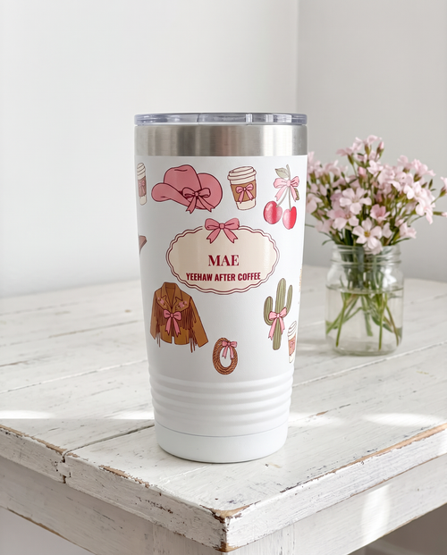 Coquette Cowgirl, Yeehaw After Coffee wrap on a white 20oz tumbler personalized MAE, YEEHAW AFTER COFFEE, three-quarter view showing the ribbed lower wall, a white-painted farmhouse table with pale pink wildflowers