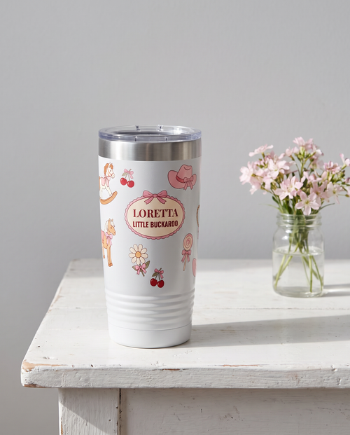 Coquette Cowgirl, Little Buckaroo wrap on a white 20oz tumbler personalized LORETTA, LITTLE BUCKAROO, three-quarter view showing the ribbed lower wall, a white-painted farmhouse table with pale pink wildflowers
