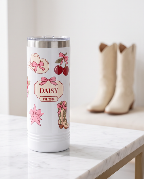 Coquette Cowgirl, Est. Year wrap on a white 22oz Skinny tumbler personalized DAISY, EST. 2004, a white marble vanity top in a bright boutique