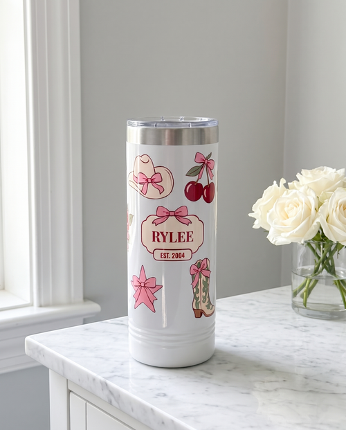 Coquette Cowgirl, Est. Year wrap on a white 22oz Skinny tumbler personalized RYLEE, EST. 2004, a white marble vanity top with a vase of white roses