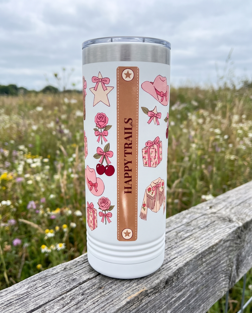 Back of a white 22oz Skinny tumbler with the Coquette Cowgirl, Spurs On Bows On wrap, the band reading HAPPY TRAILS, a weathered fence rail in a wildflower meadow
