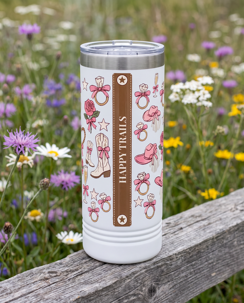 Back of a white 22oz Skinny tumbler with the Coquette Cowgirl, Final Roundup wrap, the band reading HAPPY TRAILS, a weathered fence rail in a wildflower meadow