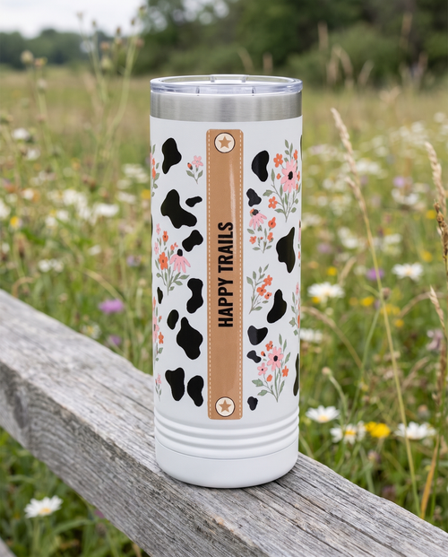 Back of a white 22oz Skinny tumbler with the Cow Print Florals, In a Moood wrap, the band reading HAPPY TRAILS, a weathered fence rail in a wildflower meadow
