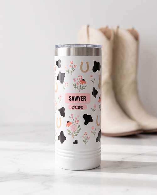 Cow Print Florals, Est. Year wrap on a white 22oz Skinny tumbler personalized SAWYER, EST. 2015, a white marble vanity top in a bright boutique