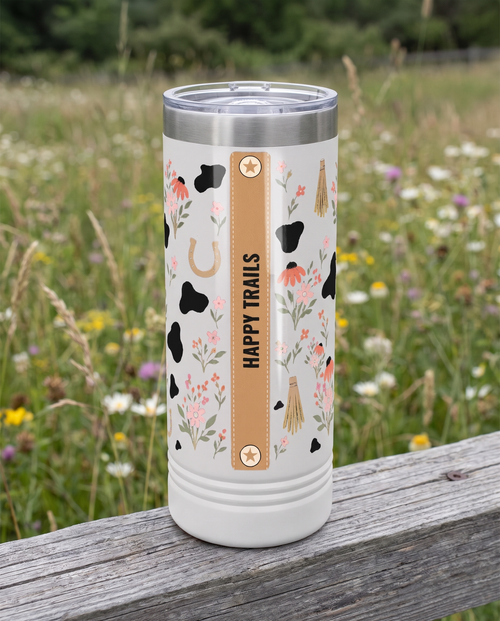 Back of a white 22oz Skinny tumbler with the Cow Print Florals, Est. Year wrap, the band reading HAPPY TRAILS, a weathered fence rail in a wildflower meadow