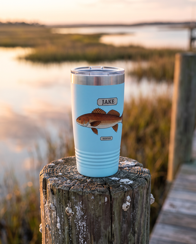 20oz Ringneck tumbler in light blue with the Field Guide Redfish design and a name plate, standing on a dock piling at sunrise