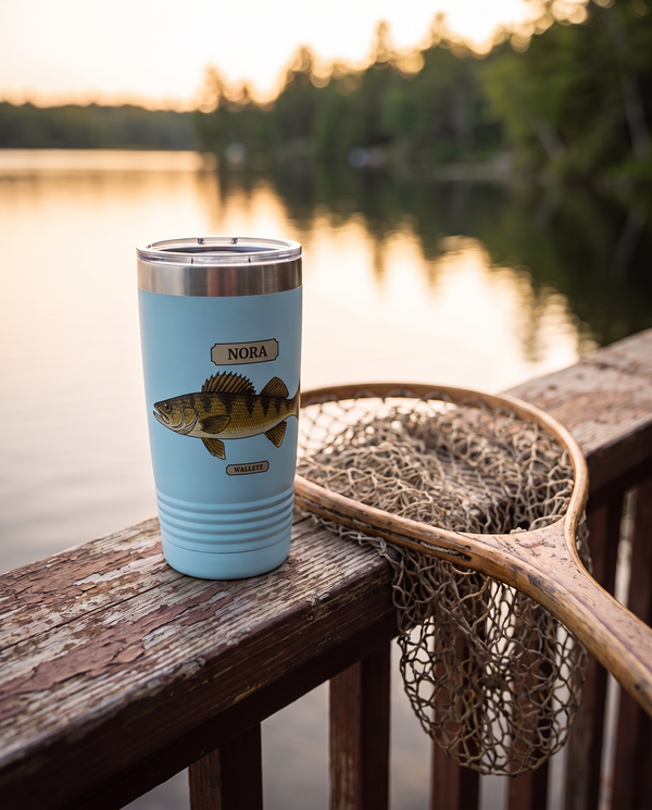 Field Guide Walleye personalized tumbler in light blue on a cabin porch rail above a lake in evening light, name NORA