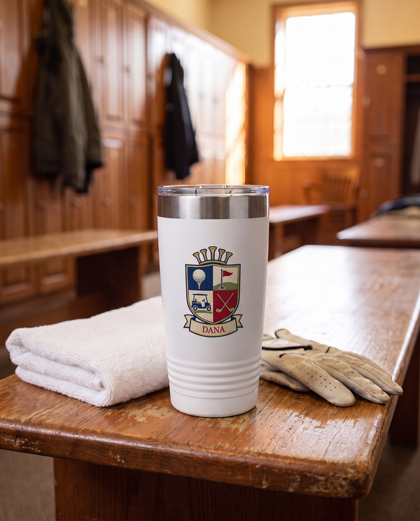 Clubhouse Shield personalized golf tumbler in white on a clubhouse locker room bench beside a towel and golf gloves, name DANA