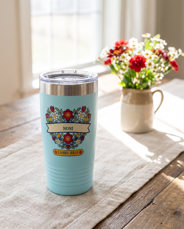 Folk Flower Heart, a folk flower heart in red, cream, gold and sage, outlined in navy, UV printed on the front of a 20oz Ringneck tumbler in Light Blue, sample name NONI, sits on a farmhouse kitchen table with a natural linen runner, a small jug of red and white wildflowers far behind by a sunny window