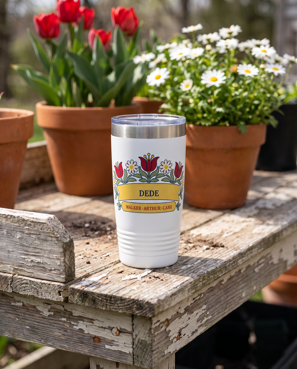 Tulip Garden Arch, folk tulips and daisies in red, cream, gold and sage, outlined in navy, UV printed on the front of a 20oz Ringneck tumbler in White, sample name DEDE, sits on a weathered wooden potting bench in a spring garden, terracotta pots of red tulips and white daisies far behind
