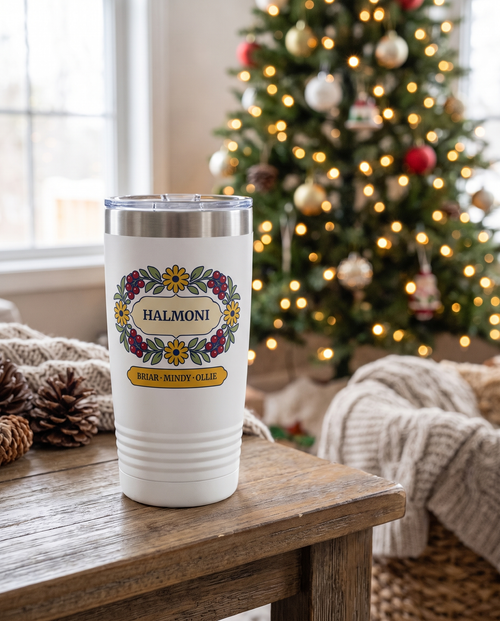 Berry Wreath, a berry and daisy wreath in red, gold, cream and sage, outlined in navy, UV printed on the front of a 20oz Ringneck tumbler in White, sample name HALMONI, sits on a wooden side table in a cosy living room at Christmas, a decorated tree with warm string lights far behind