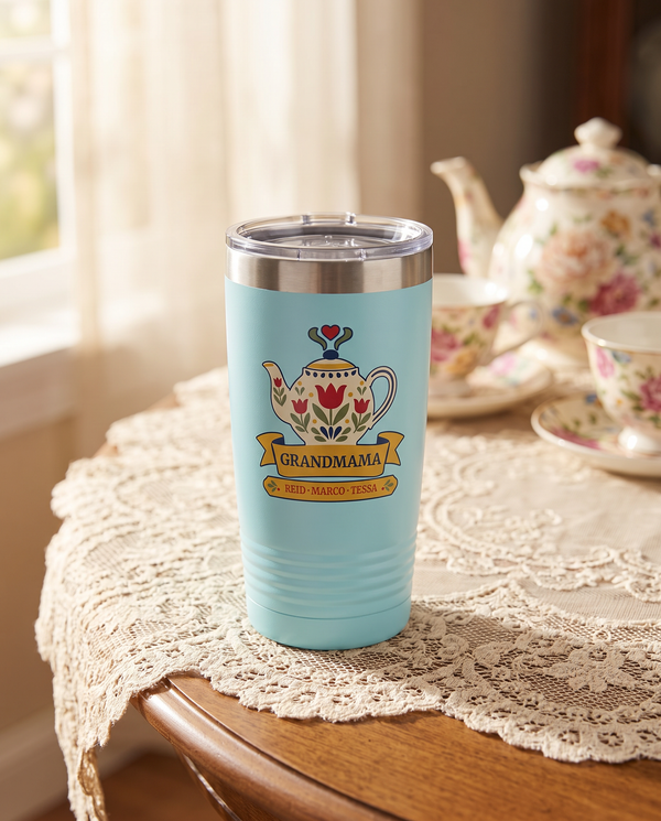 Tulip Teapot, a folk teapot in cream, red, gold and sage, outlined in navy, UV printed on the front of a 20oz Ringneck tumbler in Light Blue, sample name GRANDMAMA, sits on a tea table with a cream lace tablecloth, a floral teapot and teacups far behind, soft afternoon window light