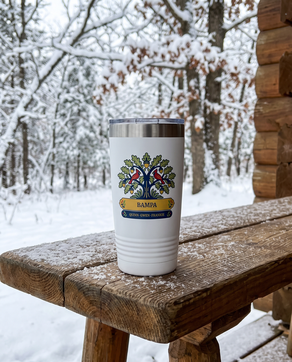 Cardinal Oak, an oak tree with cardinals in navy, red, gold and sage, UV printed on the front of a 20oz Ringneck tumbler in White, sample name BAMPA, sits on a log cabin porch table in winter, snowy oak woods far behind