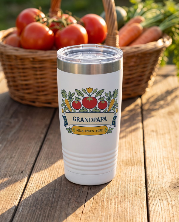 Kitchen Garden, a vegetable garden in red, gold, cream, navy and sage, UV printed on the front of a 20oz Ringneck tumbler in White, sample name GRANDPAPA, sits on a garden harvest table with a wicker basket of tomatoes and carrots far behind, morning sun