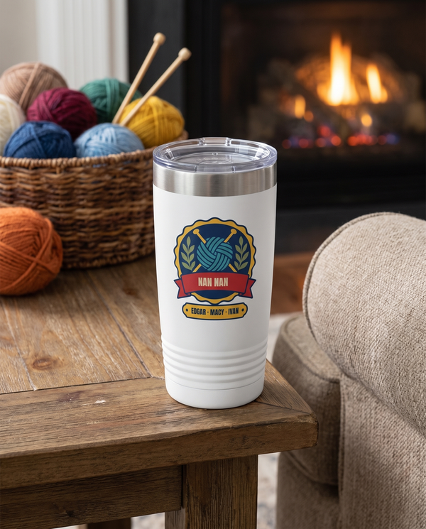 Knitting Badge, a knitting badge in navy, teal, red, gold and olive, UV printed on the front of a 20oz Ringneck tumbler in White, sample name NAN NAN, sits on a side table beside an armchair, a basket of yarn balls and knitting far behind, warm fireplace glow